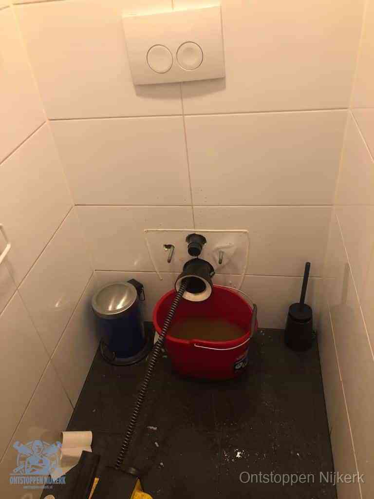 Een ontstopte wand-wc met verwijderde toiletpot waar een ontstoppingsveer in de wanduitgang verdwijnt, het achterhalen waar de verstopping zit in Nijkerk